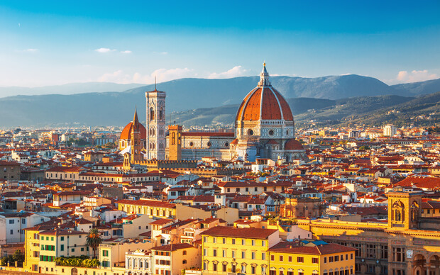 The dome of a prominent cathedral rises above a densely packed cityscape, surrounded by historic buildings. Hills form a distant backdrop under a clear blue sky.
