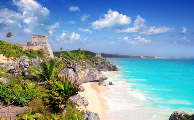 Ancient stone ruins stand on a rocky cliff overlooking a turquoise ocean. Waves gently crash on the sandy beach below, surrounded by lush greenery and clear blue skies.