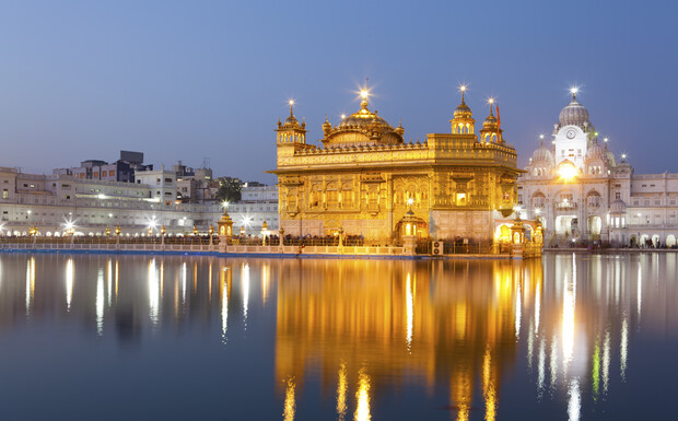 A golden temple glows brightly, reflected in a tranquil body of water, surrounded by evening-lit buildings, against a clear blue sky backdrop.