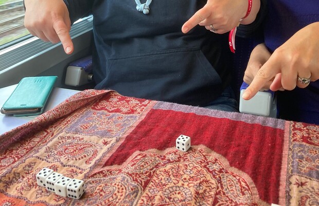 Dice are tossed on a decorative red patterned cloth, surrounded by two people pointing at them. The setting appears to be a train, with a window and seats visible.