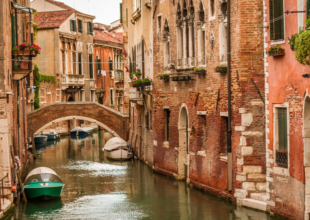 Boats float on a narrow canal, flanked by old brick buildings adorned with flower boxes and arched windows, under a small stone bridge in a charming, historic city scene.
