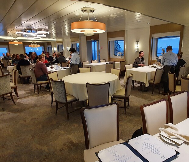 Round tables are set with white tablecloths in a spacious, elegantly lit restaurant. People dine and converse while waitstaff attend to guests. Windows reveal an ocean view outside.