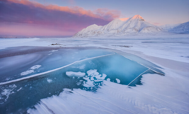 A partially frozen lake reflects a vibrant pink sunset beneath snow-covered mountains, creating a striking contrast in a vast, icy landscape.
