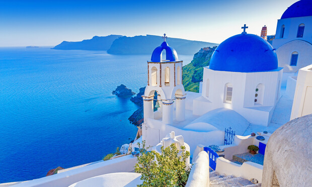 White buildings with blue domes overlook a vast, tranquil sea. The architecture stands on a cliffside under a clear sky, with a distant mountainous coastline visible in the background.