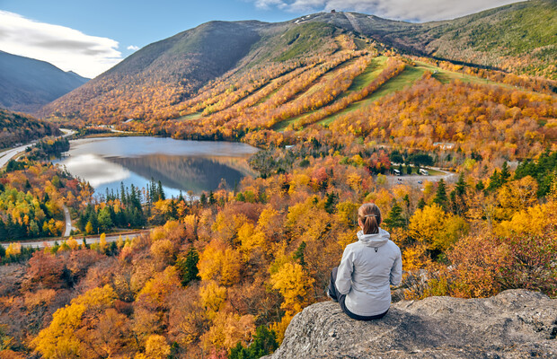 A person sits on a rock, contemplating an autumn landscape featuring vibrant trees, a reflective lake, and mountains under a partially cloudy sky.