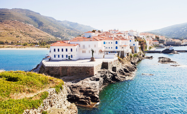 White buildings with red roofs sit atop a rocky coastline, surrounded by clear blue water. A statue stands prominently in the foreground, with mountains visible in the background.