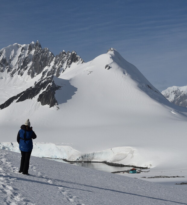 Person stands on a snowy hill, photographing a large, white mountain with jagged peaks. A small, blue structure is visible near a partially frozen body of water in the background.