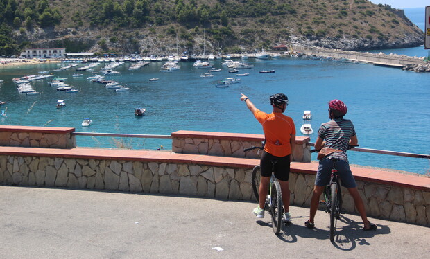 Two cyclists in helmets stand by their bikes, one pointing towards a busy marina filled with anchored boats, set against a backdrop of hilly landscape and vibrant blue sea.