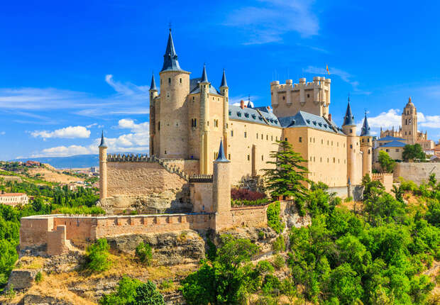 A medieval castle with pointed towers stands on a rocky hilltop. Surrounded by lush greenery and under a bright blue sky, it overlooks a distant town.