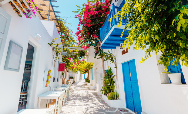Narrow alleyway adorned with vibrant pink bougainvillea, framed by white buildings and blue doors. Outdoor tables and chairs line the path, giving a Mediterranean coastal village vibe.