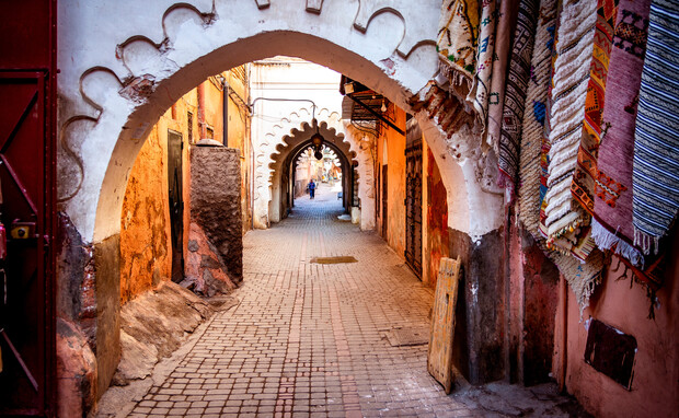 Arched stone passageway leads through narrow alley, paved with brick. Vibrant woven carpets hang on walls, creating a colorful, textured atmosphere. In the distance, a person walks away.
