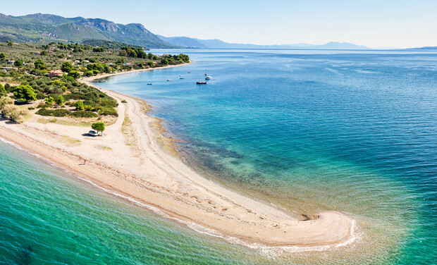 A sandy beach forms a curved peninsula, stretching into a calm, turquoise sea. Small boats float nearby, surrounded by lush green hills and distant mountains under a clear blue sky.