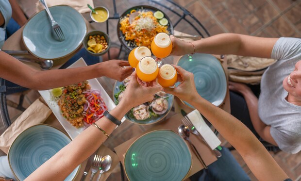 Hands raise glasses filled with orange drinks in a toast above a table set with plates of varied foods, including salads and appetizers, in a casual dining setting.