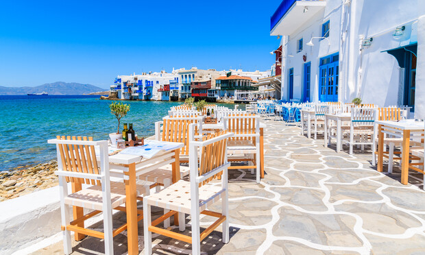 Wooden tables and chairs line a stone patio with white accents. They overlook a clear blue sea, adjacent to a row of colorful seaside buildings under a bright sky.