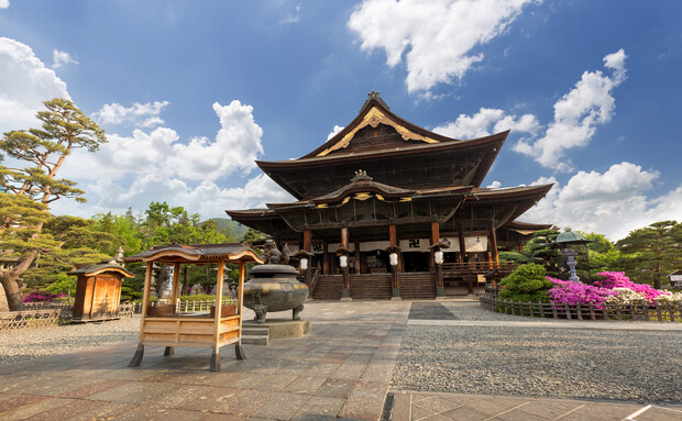 A traditional Japanese temple stands prominently under a blue sky with scattered clouds, surrounded by lush greenery and vibrant pink flowers, with a stone courtyard in the foreground.