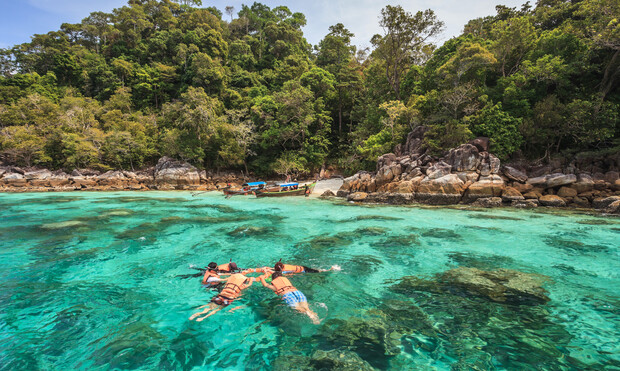 Two people float in turquoise water wearing life vests, surrounded by lush trees and rocks; a boat is moored near a sandy area on the shore.