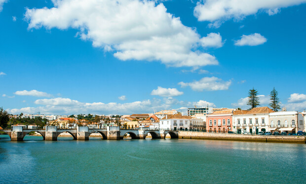 A stone bridge spans a calm river, bordered by pastel-colored buildings on a sunny day, with white clouds in a bright blue sky.