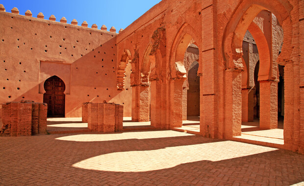 Arched stone structures stand in sunlight, casting intricate shadows on a brick courtyard. The surrounding walls feature geometric designs and a wooden door, evoking a historic architectural feel.