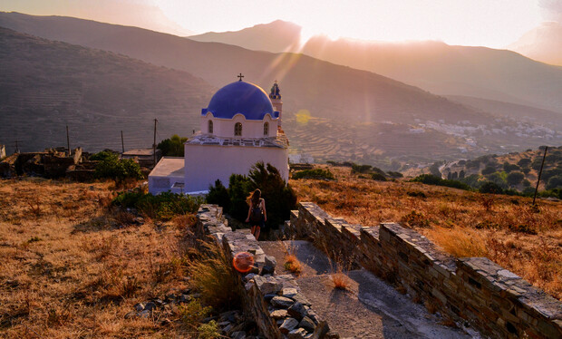 A white church with a blue dome sits calmly at sunset, surrounded by dry grass and stone walls, overlooking a mountainous landscape with scattered trees and distant hills.