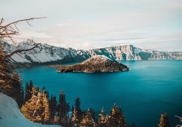 An island sits in a vast, blue lake surrounded by snow-capped mountains, with evergreen trees lining the foreground, under a clear sky.