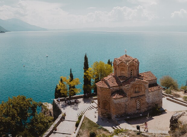 A historic stone church stands overlooking a vast, serene lake. Surrounding trees and pathways weave through the landscape, with mountains visible in the background under a partly cloudy sky.