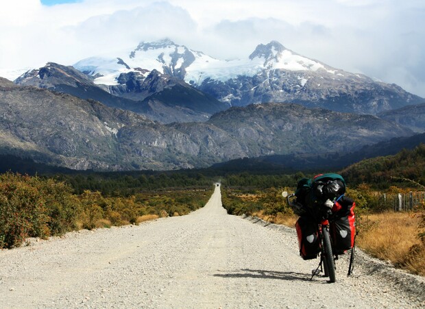 A loaded touring bicycle rests on a gravel road, stretching toward distant snow-capped mountains, surrounded by rugged hills and sparse vegetation under a cloudy sky.