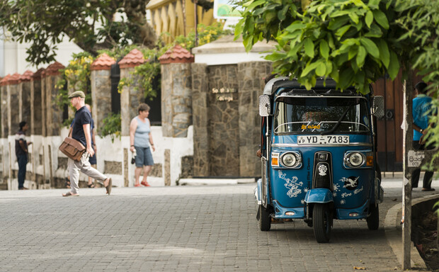 A blue tuk-tuk, parked on a tree-lined street with decorative stone walls; pedestrians walk in the background. Visible text reads "YD-7183."
