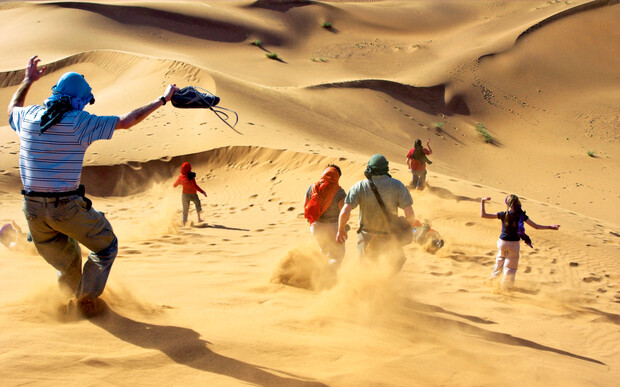 People are running down a large sand dune, creating trails of sand dust in a vast desert landscape with undulating sand dunes in the background.