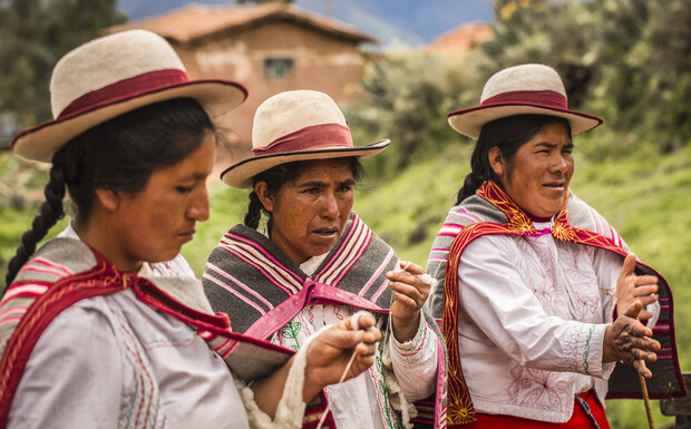 Three women, wearing traditional hats and colorful shawls, engage in a textile craft. They focus intently, surrounded by greenery and rustic houses, suggesting a rural setting.