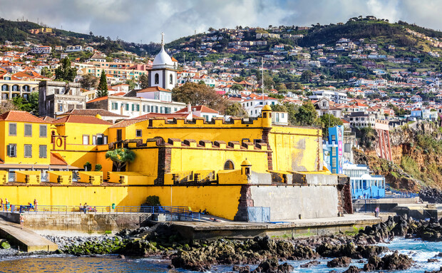 A vibrant yellow fortress with crenellations stands by the rocky coastline. In the background, a hillside is dotted with colorful buildings under a partly cloudy sky.