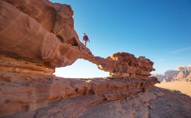 A person walking across a natural rock arch in a desert landscape, surrounded by clear blue skies and distant rocky formations.