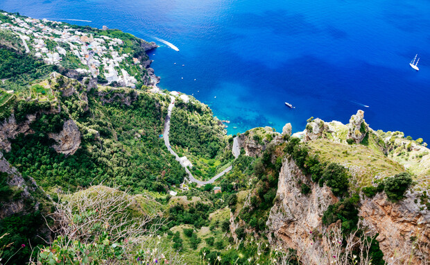 Ocean view with a rocky cliff overgrown with greenery stretching down to the vibrant blue sea. Boats sail in the water, while a winding road traverses the lush landscape.