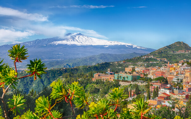 Snow-capped Mount Etna dominates the background, overlooking a colorful hillside town with Mediterranean architecture. Greenery and vibrant plants frame the foreground, creating a lush natural border.