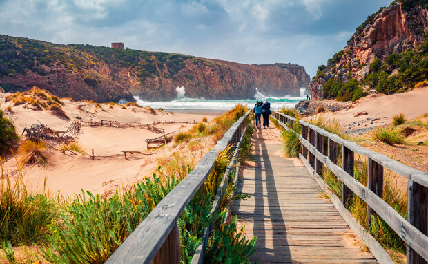 A couple walks on a wooden boardwalk through sandy dunes toward rocky cliffs and crashing ocean waves under a partly cloudy sky. Green shrubs and grasses line the path.