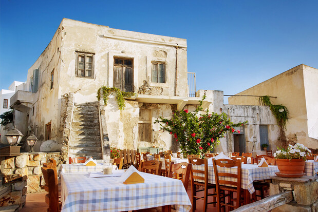 Tables with checkered tablecloths are arranged in an outdoor restaurant setting. An old, rustic building with a staircase and flowering plants serves as the backdrop under a clear blue sky.