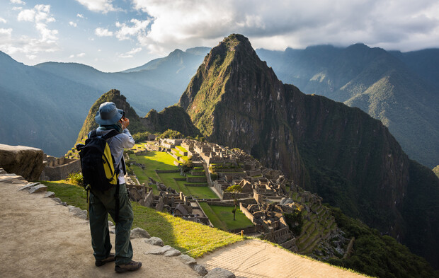 A person photographs ancient stone ruins surrounded by lush green mountains under a cloudy sky, perched on a viewing path overlooking the site.