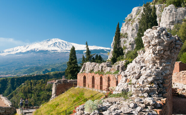 Ancient stone ruins rest on a grassy hillside with snow-capped mountains in the distance under a clear blue sky, surrounded by lush greenery and tall trees.