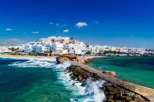 A stone walkway extends into turquoise water, leading toward a coastal town with white buildings under a clear blue sky. Waves crash against the path in a scenic Mediterranean setting.