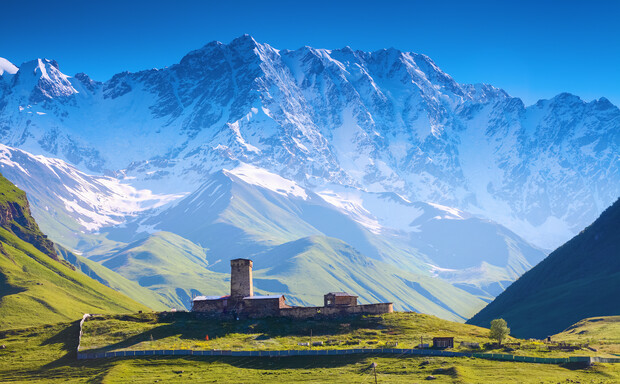 Stone tower and buildings sit on a grassy hill, surrounded by a fence, against a backdrop of towering, snow-capped mountains under a clear blue sky.