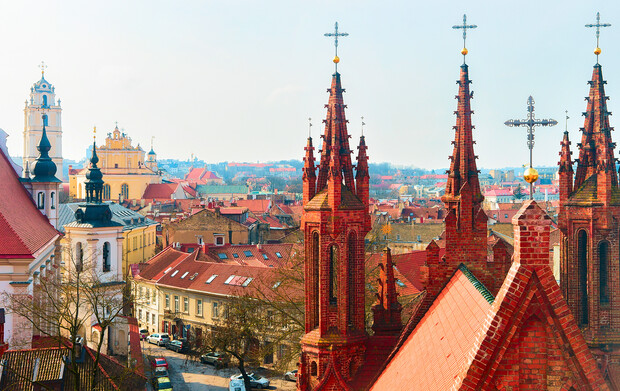 Red brick church towers rise in a historic cityscape, surrounded by smaller, colorful buildings under a clear blue sky. Crosses adorn the spires, enhancing the architectural skyline.