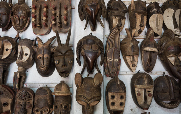 Wooden masks are displayed on a white wall, showcasing various carvings with intricate designs and diverse facial expressions, some with animal motifs and textured surfaces.