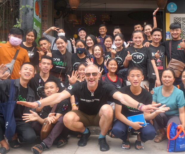 A group of people, including one person in front extending arms, pose together smiling outside a building. Many wear black shirts with logos. Lush greenery and a fruit sign are visible.