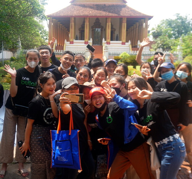 A group of smiling people takes a selfie in front of a traditional, ornate building with columns and a red roof, surrounded by greenery. Some wear matching black shirts with "one buffalo."