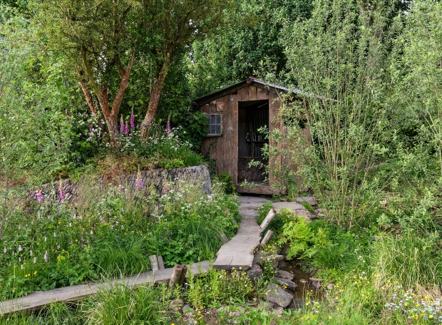 A wooden shed stands quietly surrounded by lush greenery and wildflowers, accessed by a stone pathway over a small stream, in a serene garden setting.