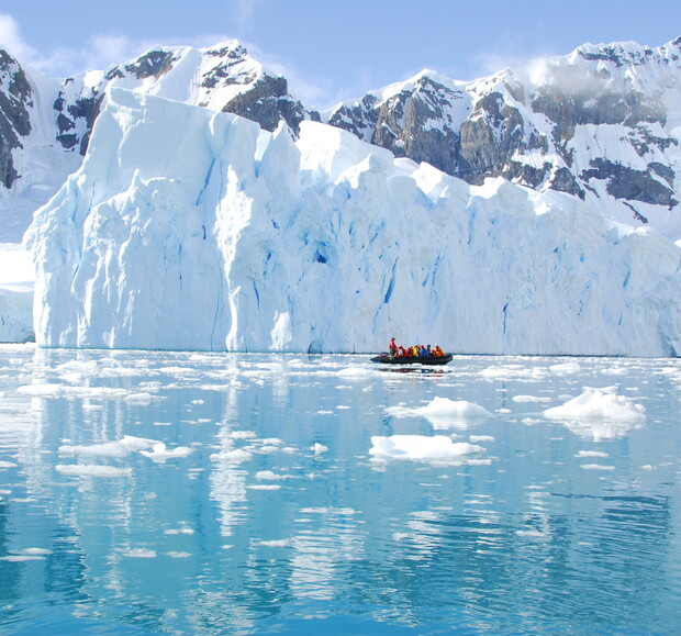 A small boat carrying people floats on icy blue water, surrounded by scattered ice chunks, against a backdrop of towering, rugged, snow-covered cliffs and glaciers.