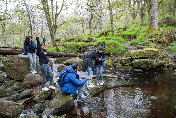 Five people interact on mossy rocks near a small stream. Two sit, while others balance or help each other across the water. Surrounding trees and greenery create a serene forest setting.