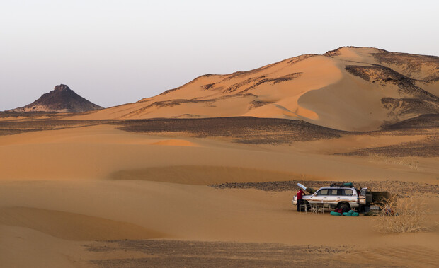 A car with an open hood and camping gear is parked on sandy dunes. A person appears near the vehicle, and distant mountains rise against a clear sky.
