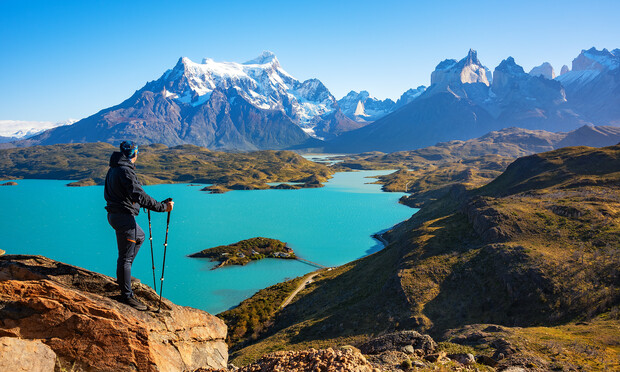 A hiker stands with trekking poles on a rocky cliff, overlooking a turquoise lake surrounded by rugged mountains under a clear blue sky. Snow-capped peaks tower in the distance.