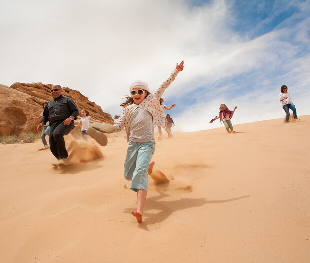 Children and an adult run joyfully down a sandy hill, with a clear blue sky and rocky formations in the background.
