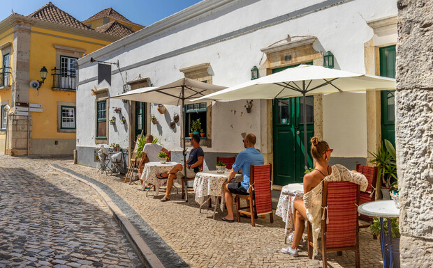 People sit at outdoor cafe tables, shaded by umbrellas, on a cobblestone street in a sunny, historic town. The building has white walls and green doors, with decorative plants.
